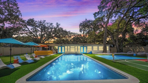Una piscina con zona delimitada por una red, rodeada de césped artificial, sillas de descanso y sombrillas.