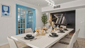 Un comedor moderno con una larga mesa dispuesta para una comida, una puerta azul y una cocina visible al fondo.