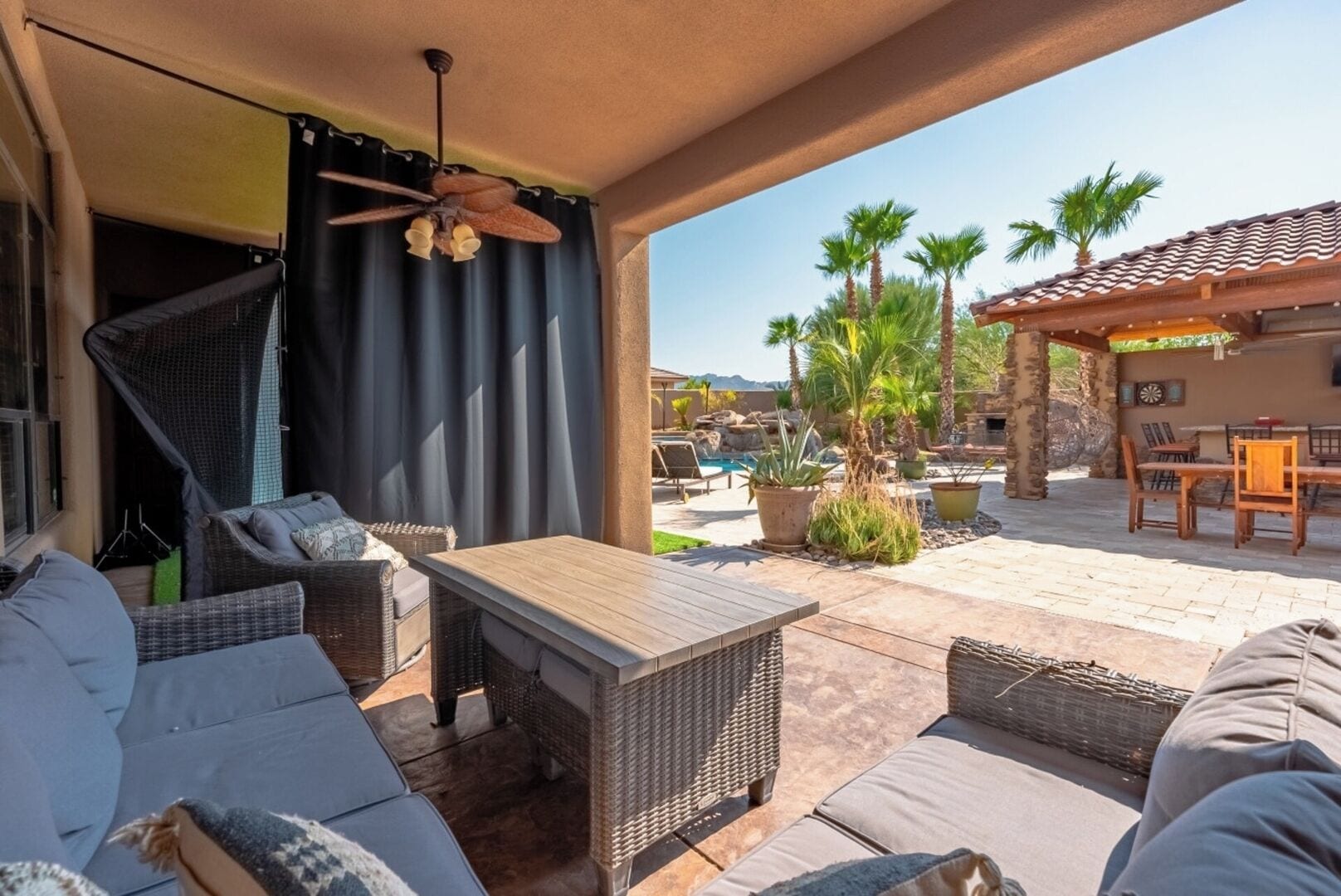 A covered outdoor seating area with wicker furniture, a wooden table, and a ceiling fan.