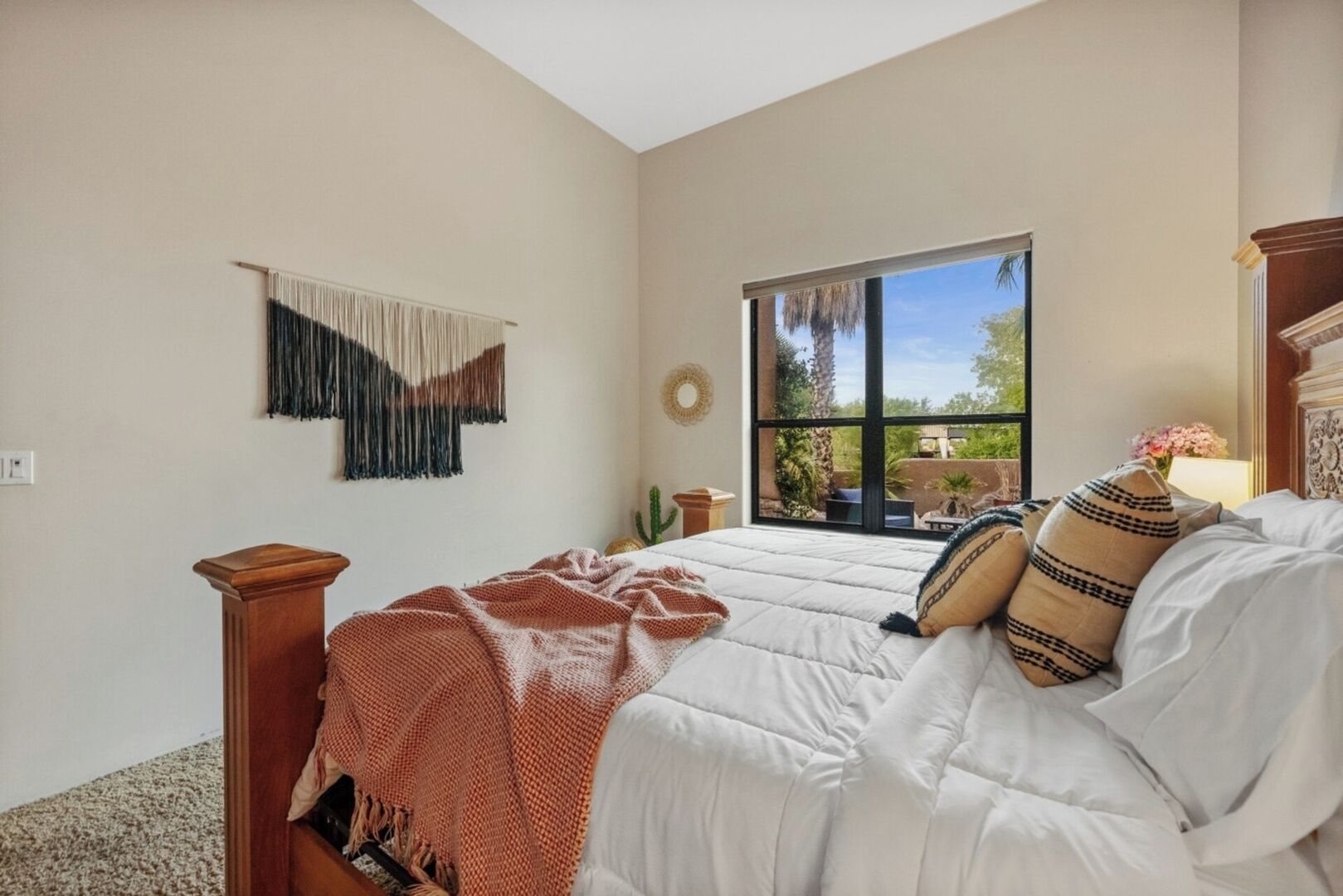 Ein Schlafzimmer mit einem großen Bett, einem Fenster mit Blick ins Grüne, einer Wanddekoration aus Makramee und einem Nachttisch aus Holz.