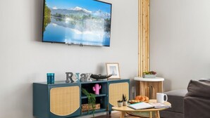 A living room with a teal cabinet, a small round table with a coffee cup, and a flat-screen TV displaying a mountain landscape. - Sleeps 25 • Heated Pool• In+Outdoor Games •Theater (Tampa)