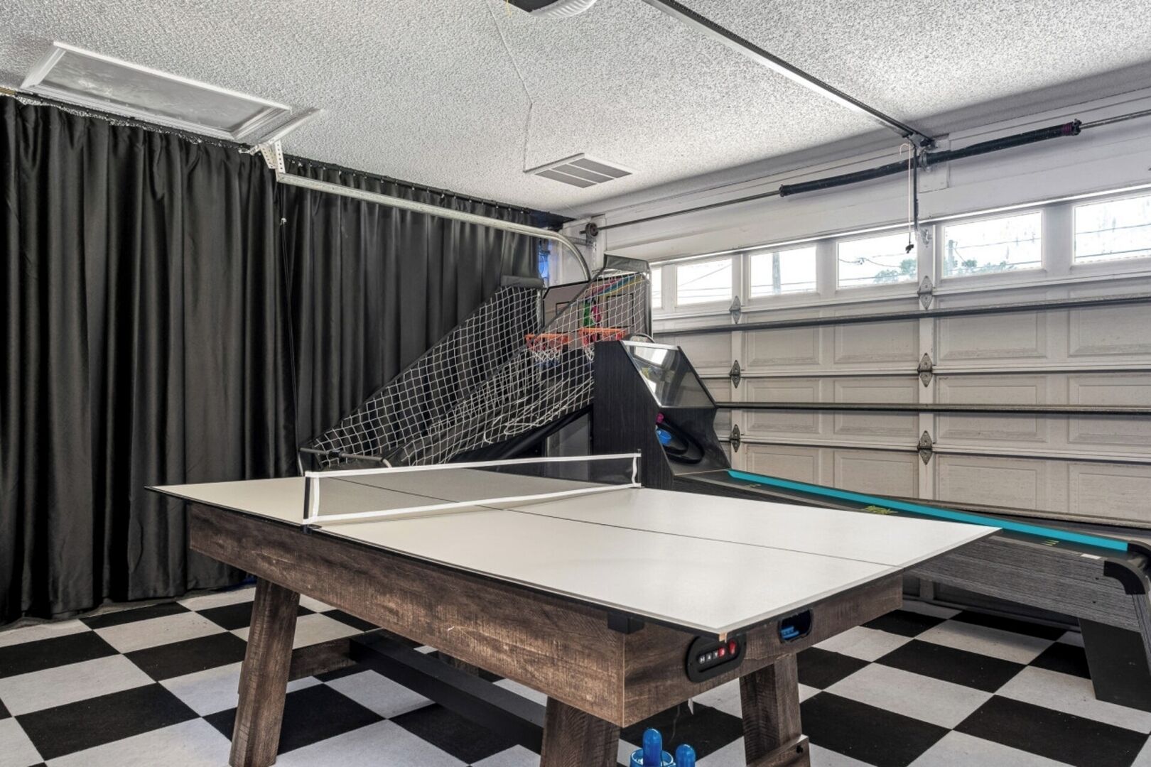 Une table de ping-pong dans une pièce dont le sol est à carreaux, avec un filet et une porte de garage en arrière-plan.