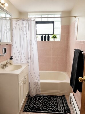 Une salle de bain avec une baignoire, un rideau de douche, un lavabo et une fenêtre sur le rebord de laquelle sont posées des bouteilles.