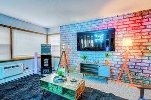 Una habitación con pared de ladrillo, un televisor de pantalla plana sobre un escritorio de madera, una lámpara de pie y una consola de videojuegos.