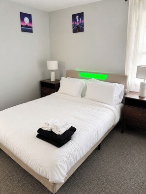 A neatly made bed with white linens and folded towels, a headboard with a green light, two bedside tables with lamps, and two wall art pieces. - 90s Condo| King Bed | 65” Smart TV | Arcade Game (Elmwood Park)