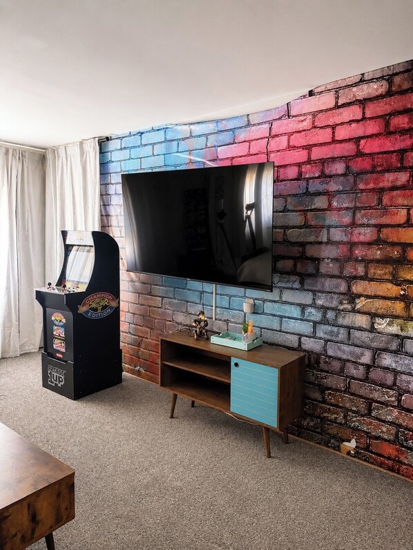 Une pièce avec un mur en briques, un téléviseur à écran plat fixé au-dessus d’un meuble de rangement en bois, et une borne d’arcade ancienne.