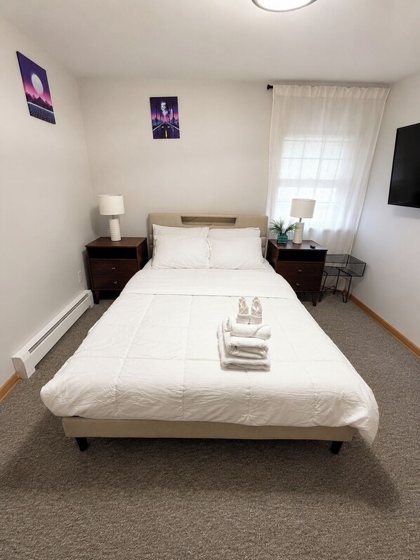 A neatly made bed with white linens, a bedside table with a lamp, a small plant, and a wall-mounted TV. - 90s Condo| King Bed | 65” Smart TV | Arcade Game (Elmwood Park)