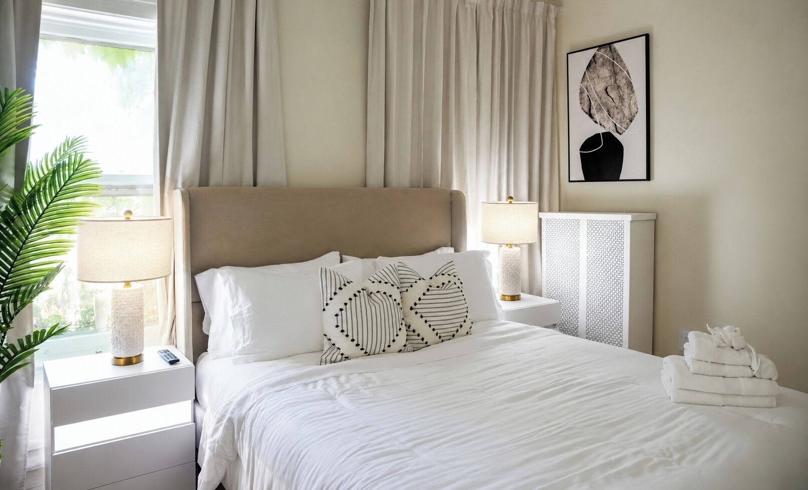 Un lit bien fait, avec du linge de lit blanc, une tête de lit beige et des coussins décoratifs. On y trouve deux lampes de chevet, une œuvre d’art accrochée au mur et une fenêtre avec des rideaux.