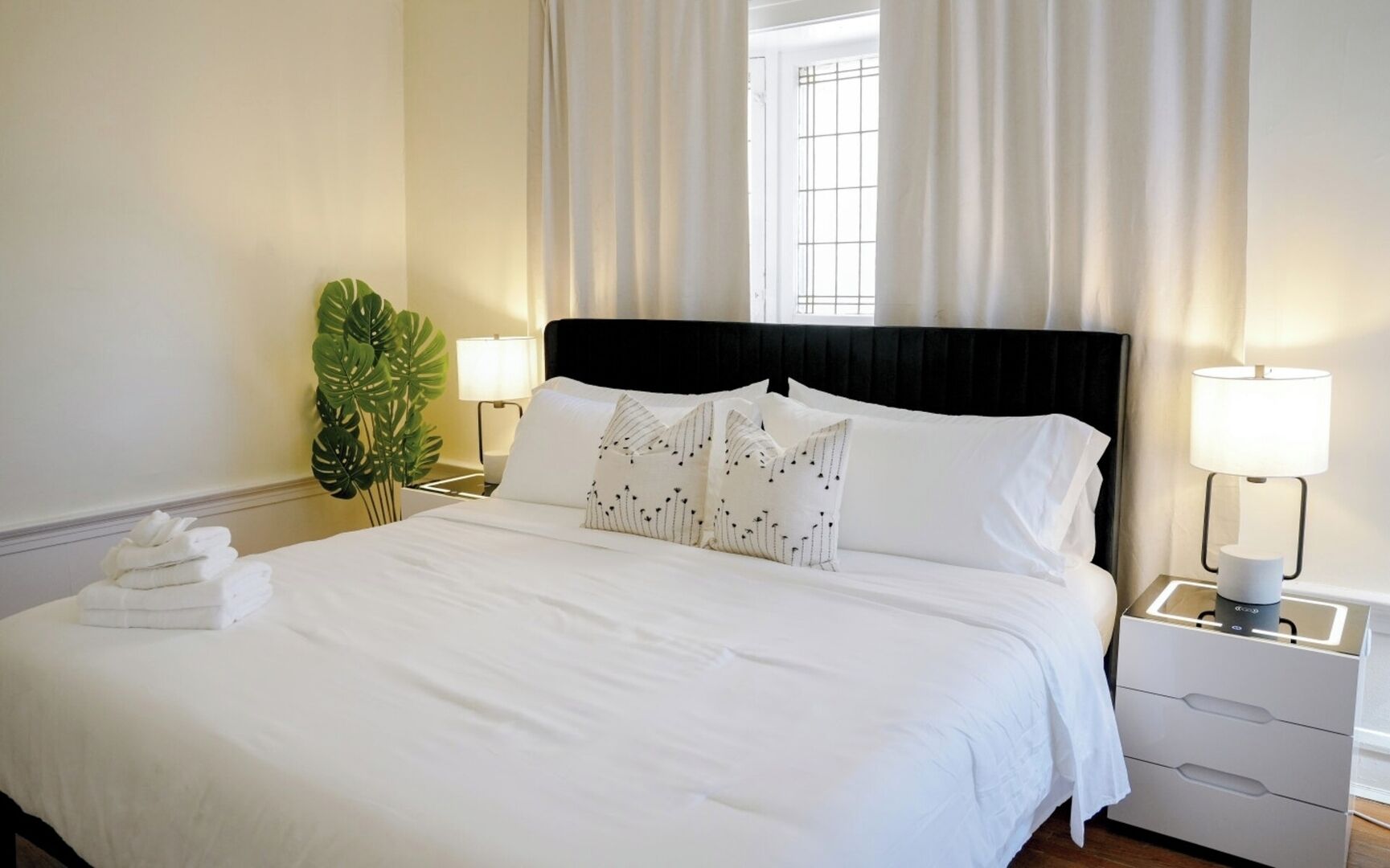 A neatly made bed with white linens, two bedside lamps, a potted plant, and a window with curtains.