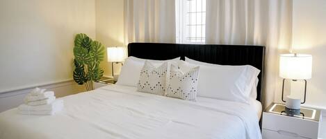 A neatly made bed with white linens, two bedside lamps, a potted plant, and a window with curtains.