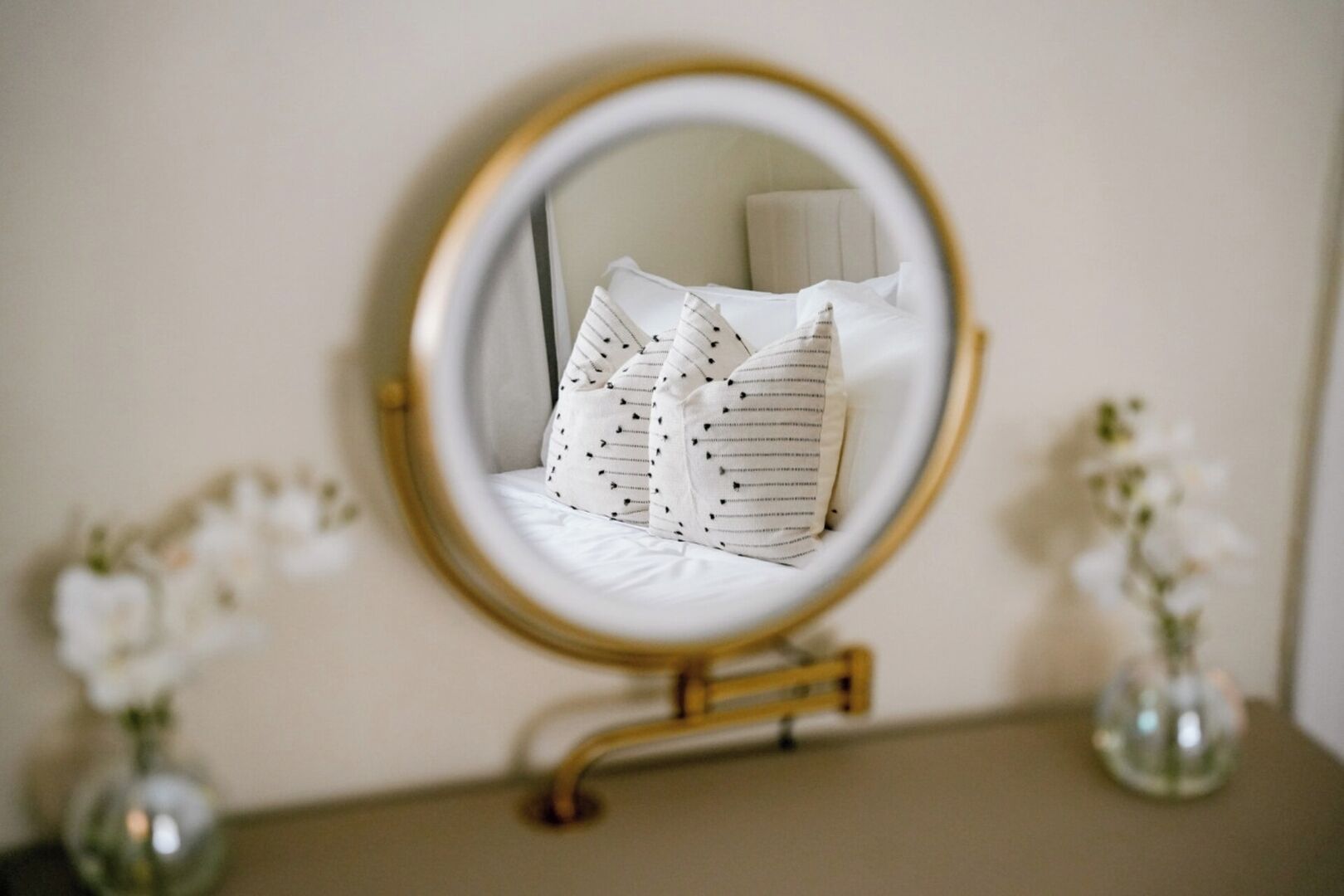 A round mirror with a gold frame on a dresser, reflecting a bed with striped pillows.