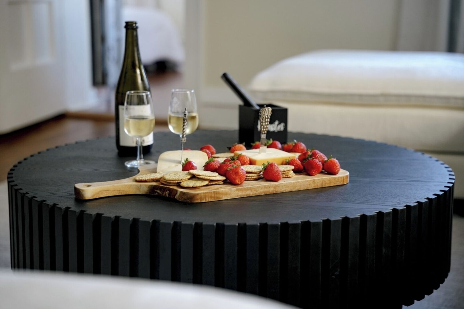 A round black table with a wooden cutting board holding cheese, strawberries, and crackers. A bottle of white wine and glasses are on the table.