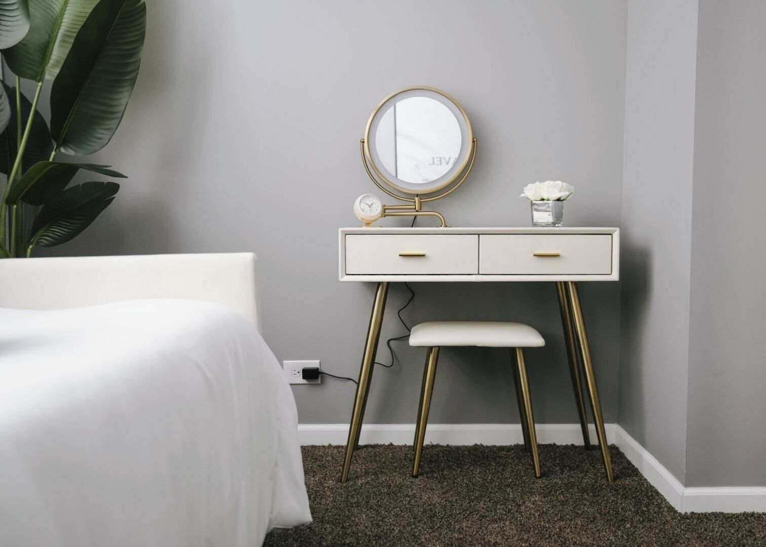 A bedroom with a white bed, a white dresser with a gold mirror, a small white vase, and a plant.