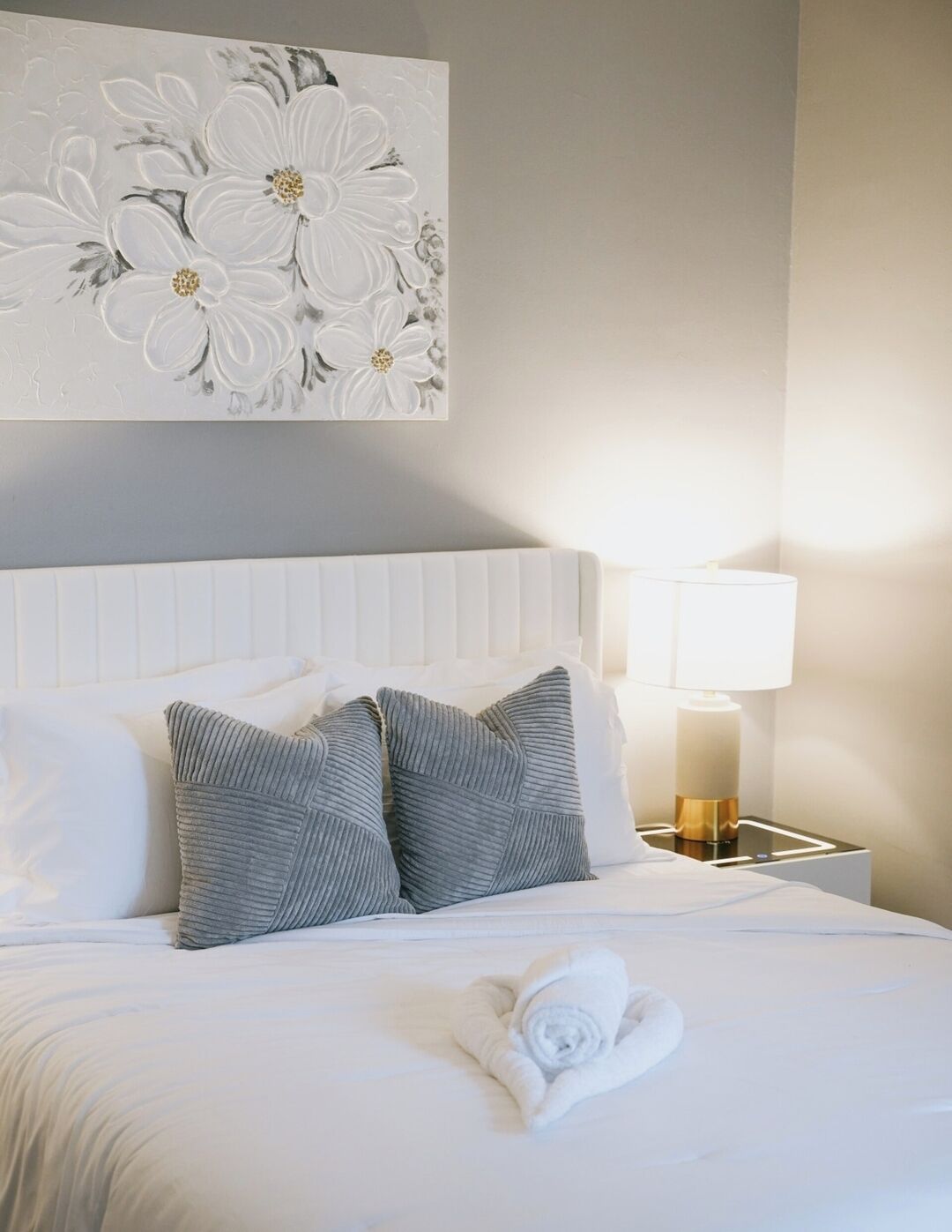 A neatly made bed with white linens and two grey pillows, a white wall with a floral artwork, and a bedside table with a lamp.