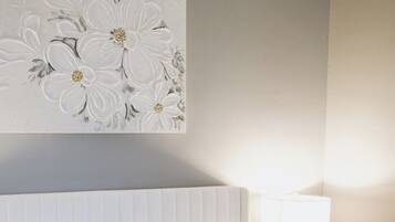 A neatly made bed with white linens and two grey pillows, a white wall with a floral artwork, and a bedside table with a lamp.