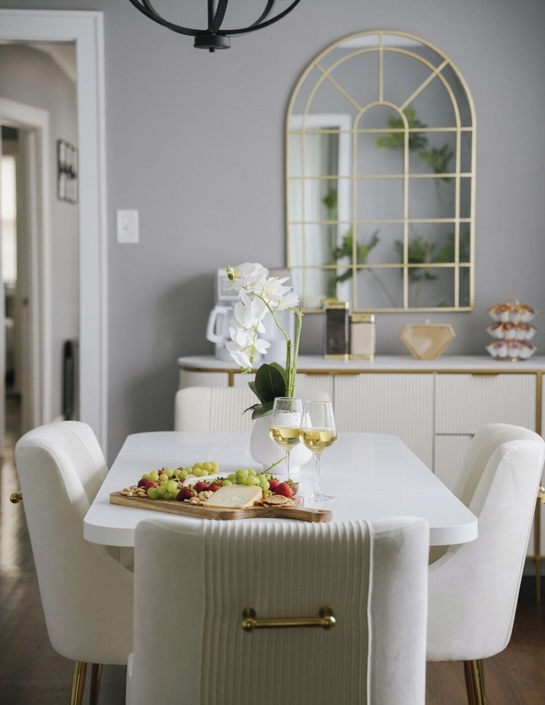 A dining table with a white tablecloth, white chairs with gold handles, and a tray with grapes, strawberries, and cheese. There are two glasses of white wine on the table.