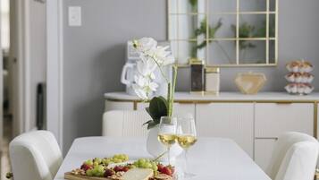A dining table with a white tablecloth, white chairs with gold handles, and a tray with grapes, strawberries, and cheese. There are two glasses of white wine on the table.