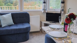 A living room with a blue sofa, a TV, and a dining table set with plates, cutlery, and a vase of flowers. - Avalon (Lyme Regis)
