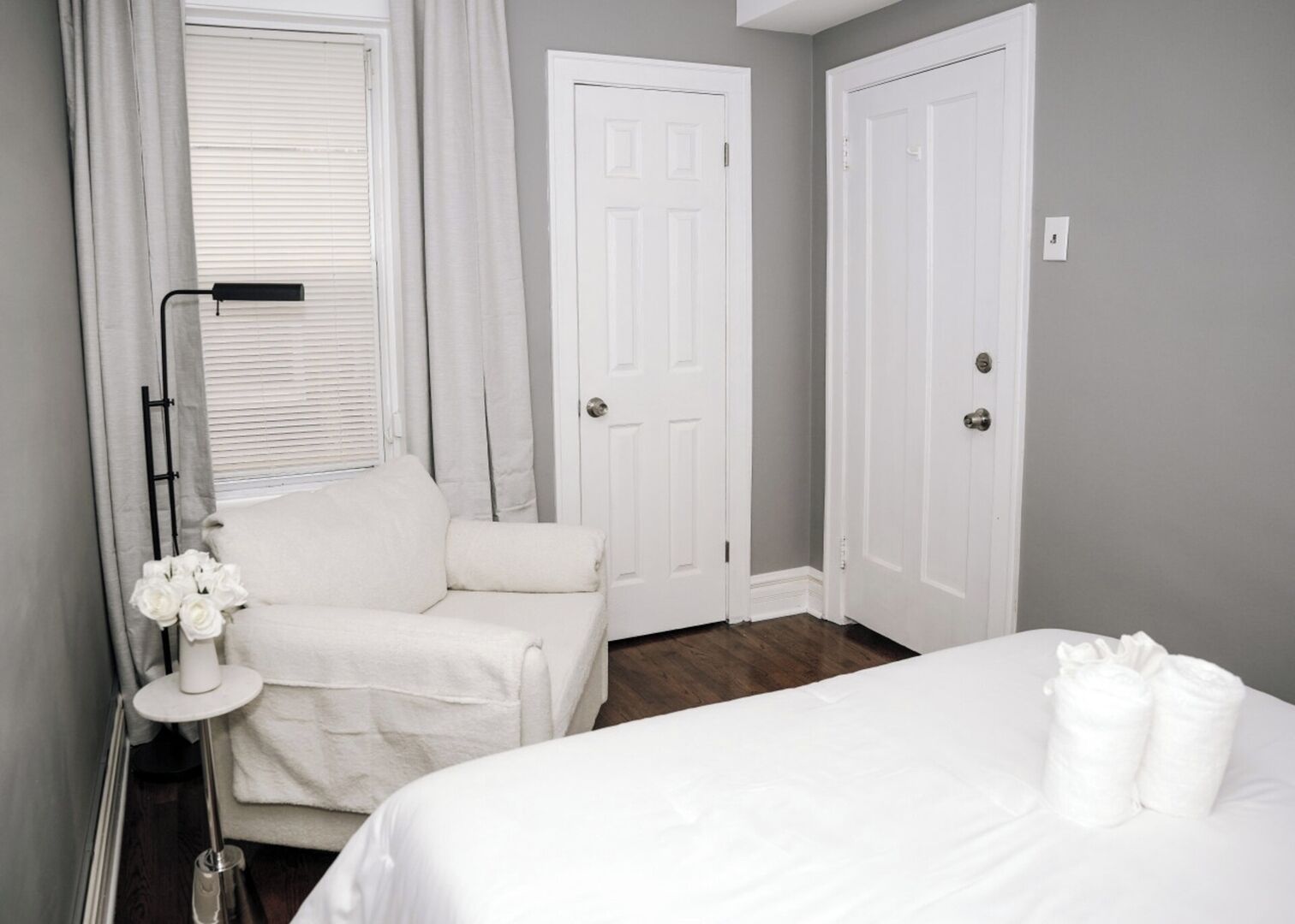 A hotel room with a bed, a chair, a desk with a lamp, and two doors.
