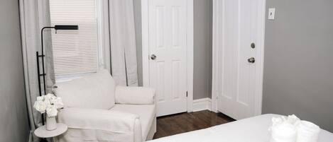 A hotel room with a bed, a chair, a desk with a lamp, and two doors.