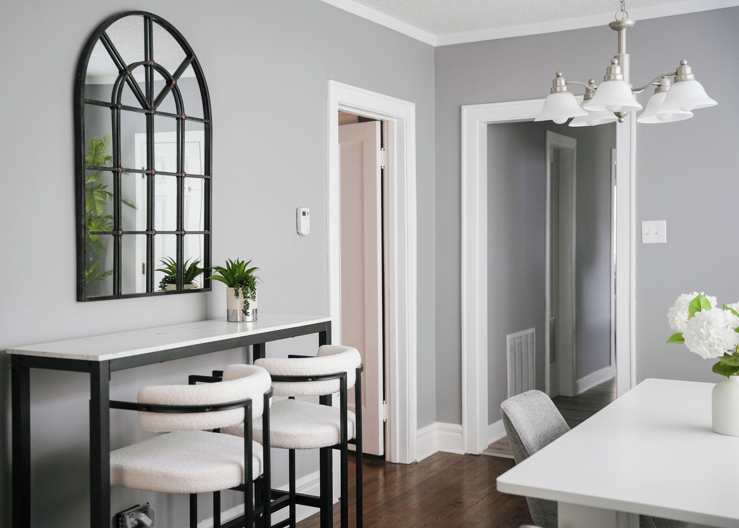 A modern dining area with a white table, three bar stools, a pendant light, and a mirror with a geometric design.