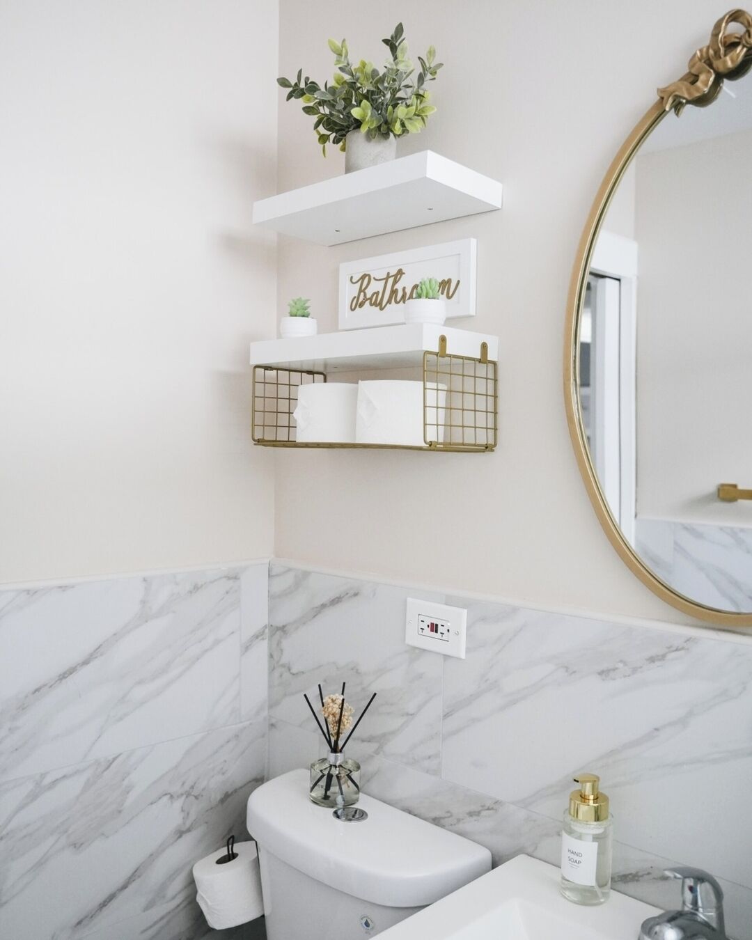 A bathroom with a marble wall, a toilet, a sink with a gold faucet, a mirror with a gold frame, and a shelf with a plant and a sign that says "Bathroom".
