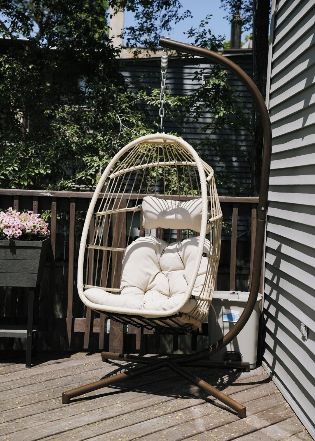 Una silla colgante de mimbre con asiento y respaldo acolchados, suspendida por una estructura metálica, sobre una terraza de madera.