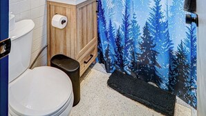 A bathroom with a white toilet, a wooden vanity, a mirror above the sink, and a shower curtain with a forest design. - King Bed Studio | 60" HDTV | Safe | Close to Metra (Elmwood Park)