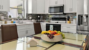 A modern kitchen with white cabinets, a glass-top dining table, and a fruit bowl. - HeatedPool • HotTub • OutdoorKitchen/Theater/Games (Bradenton)