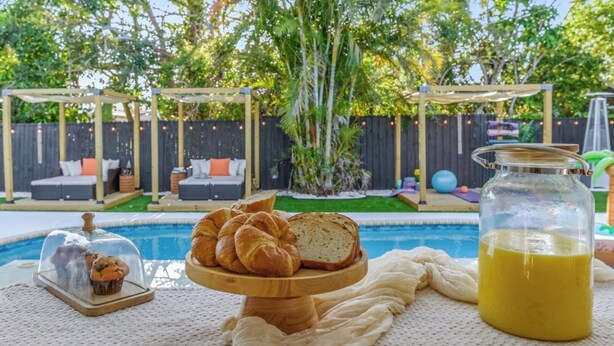 A poolside setting with a wooden serving board of bread and muffins, a glass jar of lemonade, and a clear glass dome over a muffin. - HeatedPool • HotTub • OutdoorKitchen/Theater/Games (Bradenton)