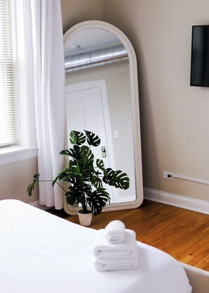 A bedroom with a large mirror, a potted plant, a neatly made bed, and a white wall with a power outlet. - Private Workspace • Sleeps 4 New Modern 1BR • (Chicago)