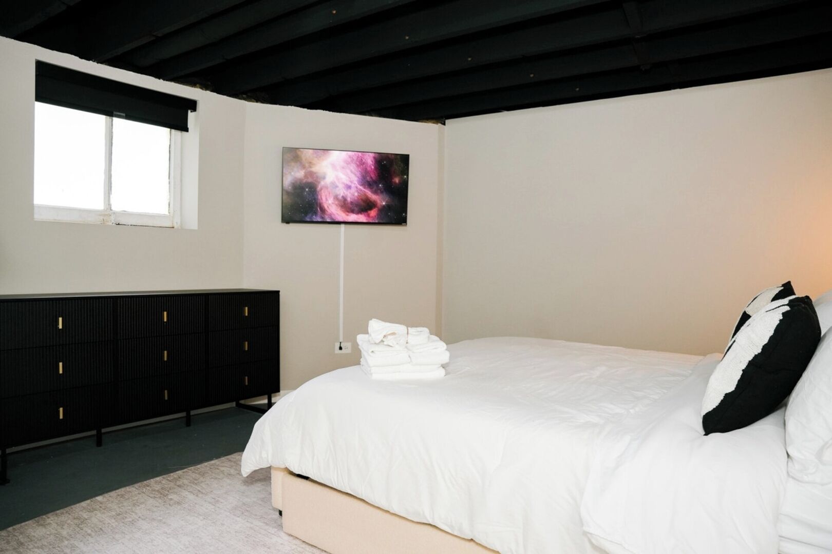 A bedroom with a bed, a black dresser, a wall-mounted TV, and a window.