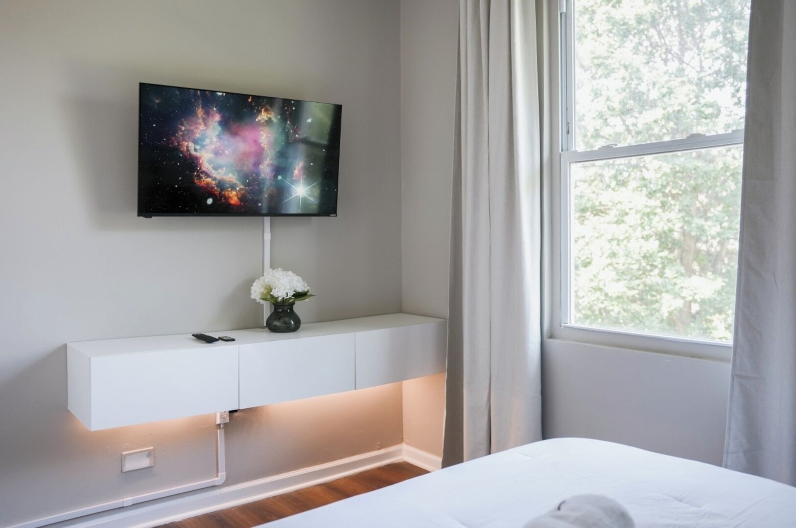A bedroom with a bed, a TV mounted on a wall, a white console table with a vase of flowers, and a window with curtains.