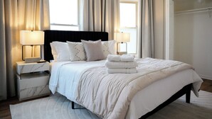 Un dormitorio ordenado con una cama grande, dos mesitas de noche y una mesita auxiliar. Hay cortinas en las ventanas y una toalla sobre la cama.
