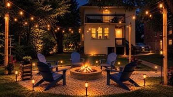 A cozy outdoor seating area with Adirondack chairs around a fire pit, string lights overhead, and a house in the background.