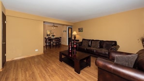 A living room with a dark brown leather sofa, a wooden coffee table, and a dining area with a chandelier.