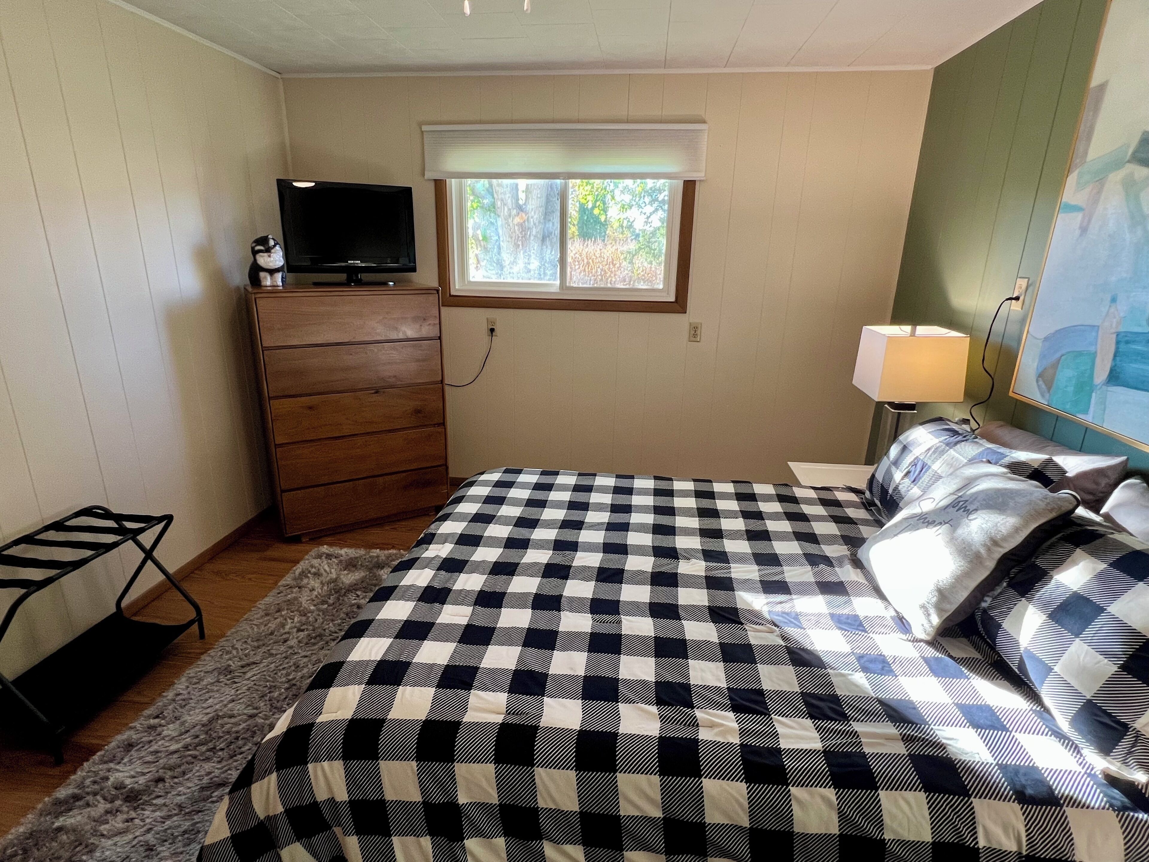A bedroom with a bed, dresser, television, window, lamp, and artwork on the wall.