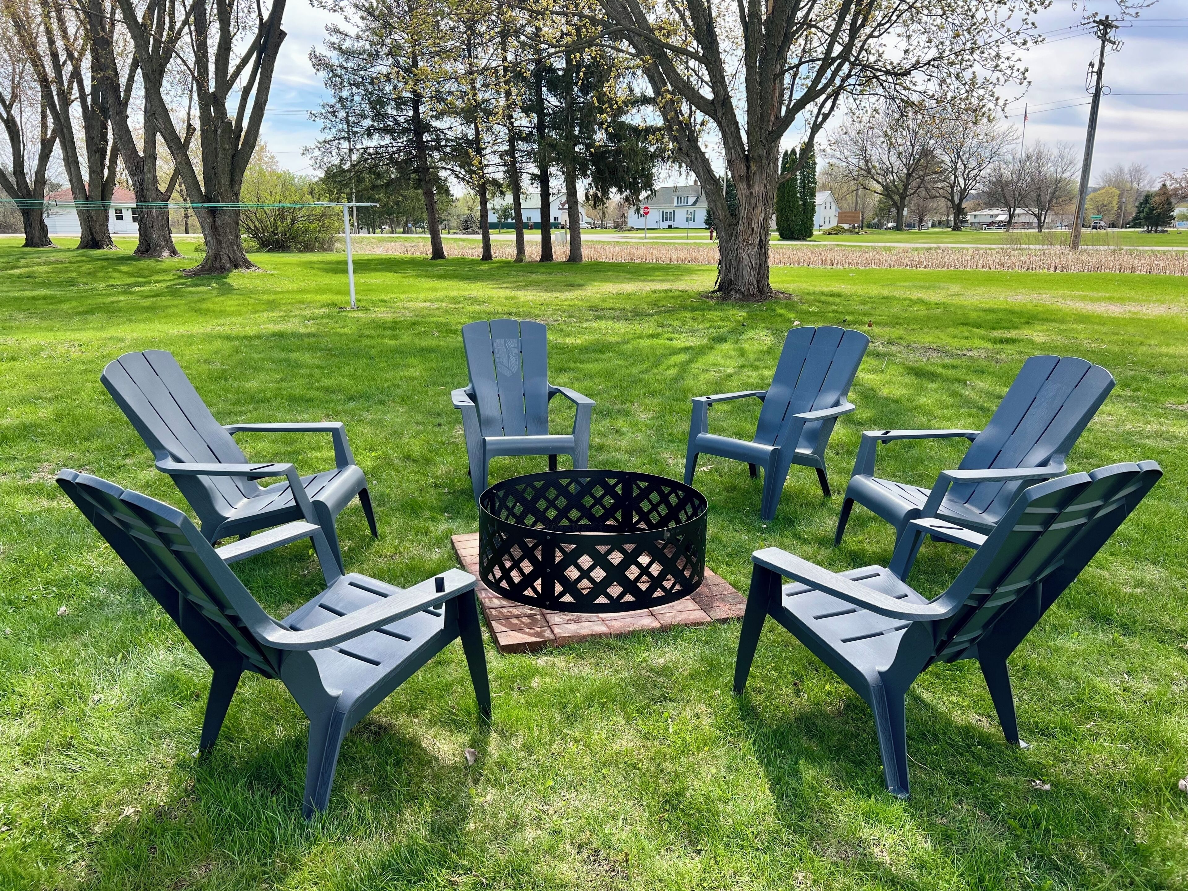 A grassy outdoor area with four Adirondack chairs arranged around a central fire pit.