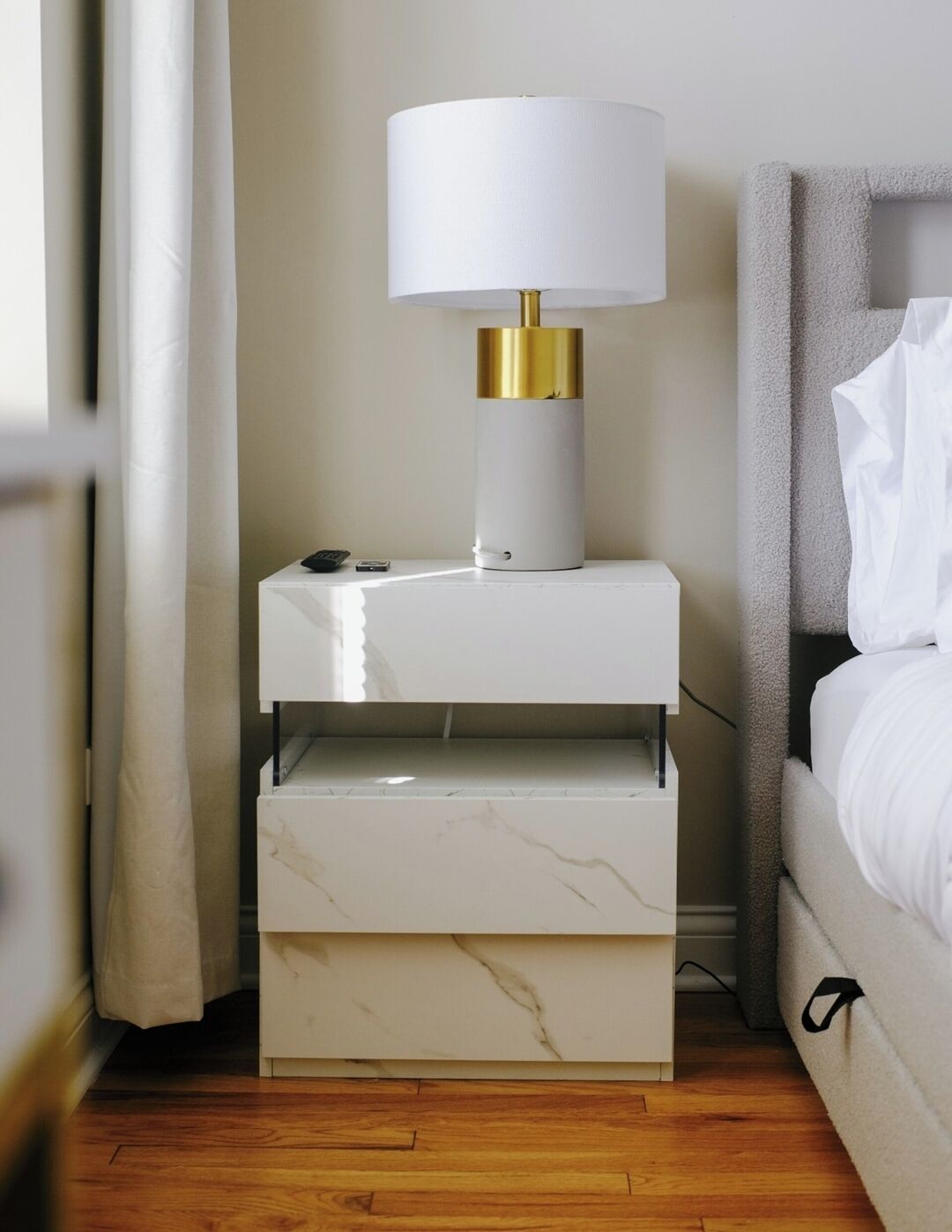 A modern hotel room with a nightstand featuring a white marble drawer, a gold and white lamp, and a small electronic device.