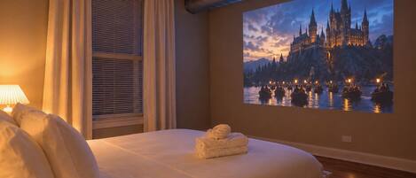 A hotel room with a large bed, a window with blinds, a projector screen displaying a castle, and a neatly made bed with towels.