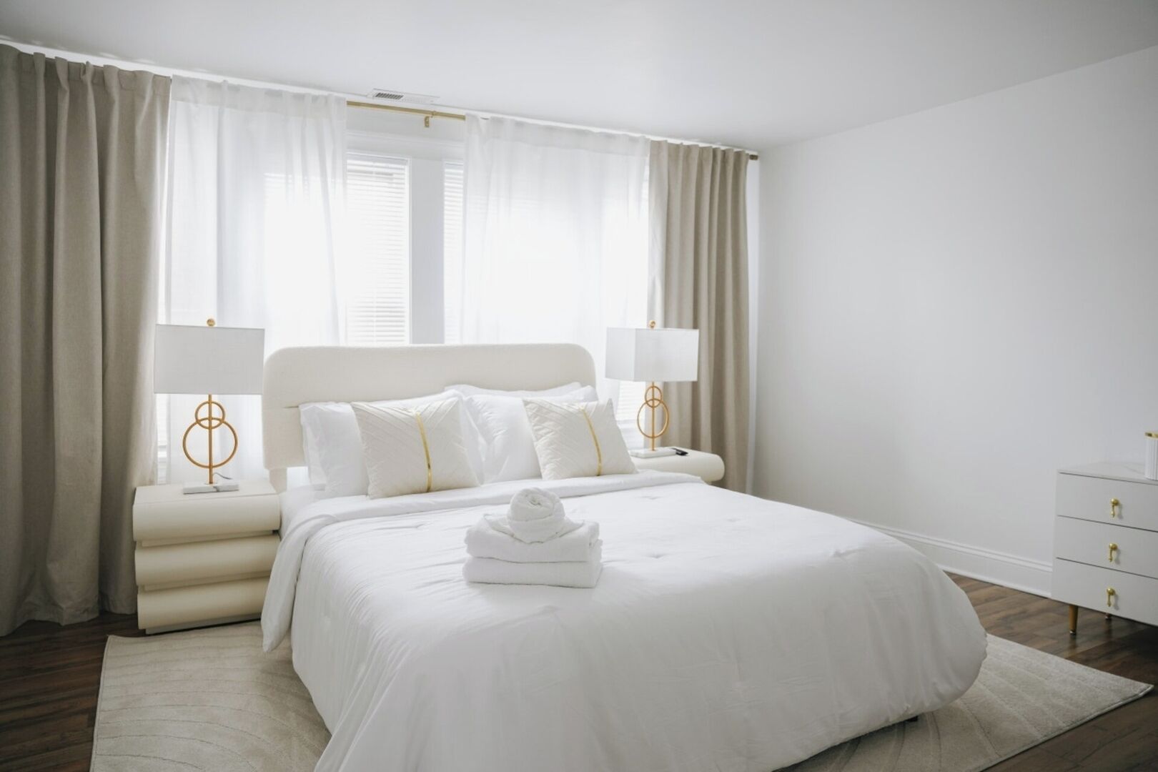 Une chambre d’hôtel moderne avec un grand lit, du linge de lit blanc et un lit parfaitement fait avec des serviettes. On y trouve deux tables de chevet avec des lampes, ainsi qu’une petite commode avec des tiroirs.