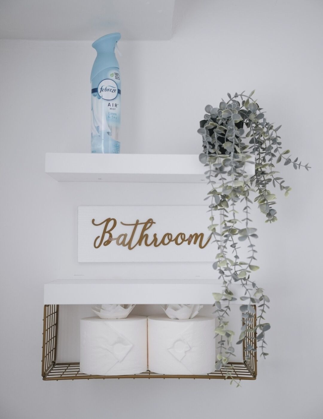 En hylla i badrummet med en Febreze-doftspridare, en skylt med texten ”Badrum” och en hängande växt.