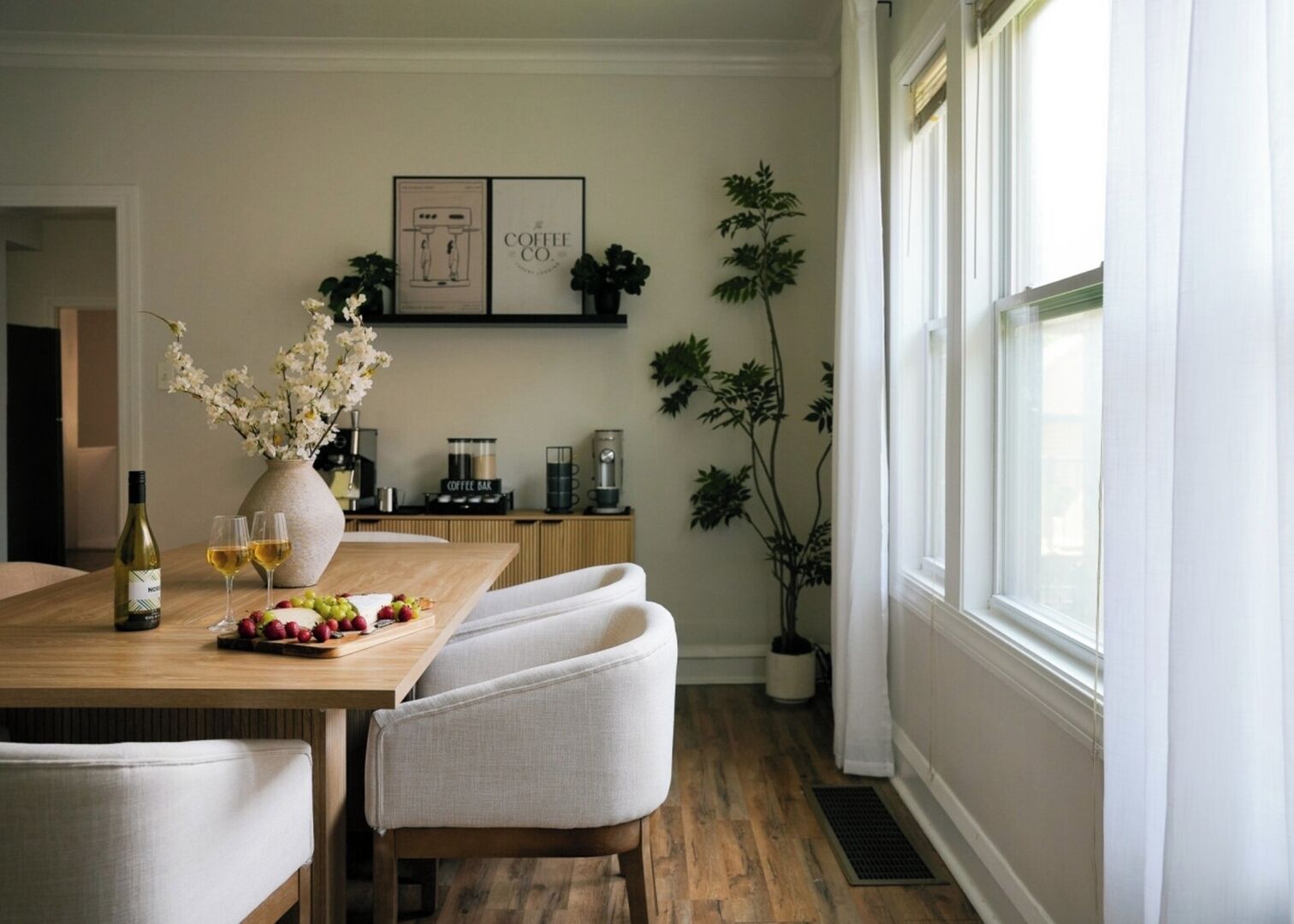 A dining area with a wooden table, chairs, a vase with flowers, a bottle of wine, and a coffee machine.