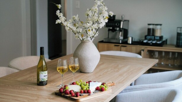 Une table à manger avec une bouteille de vin, deux verres, un vase de fleurs blanches et une planche à fromages accompagnée de fruits.