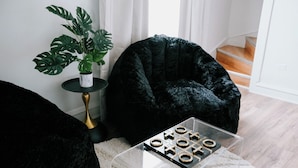 Une pièce avec un fauteuil en forme de sac en tissu noir, une petite table sur laquelle est posée une plante, et un jeu de morpion sur une table en verre transparent.