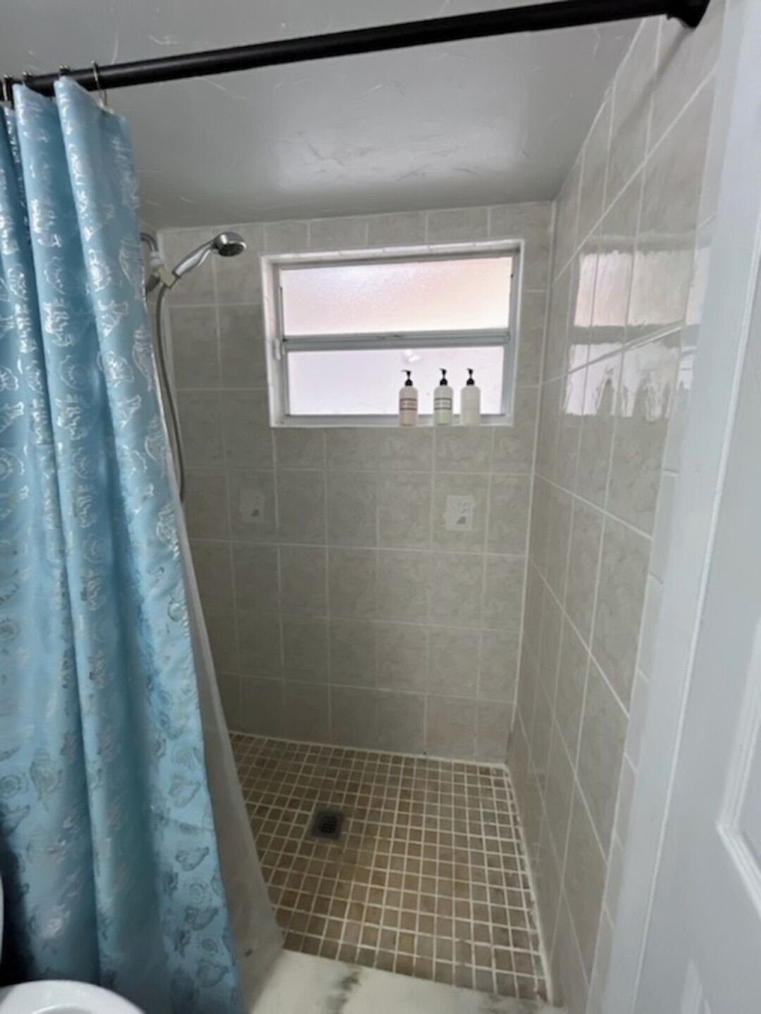 A small, tiled shower area with a window above the sink, a showerhead, and a drain.