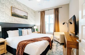 Ein Hotelzimmer mit einem großen Bett, einem Schreibtisch mit Fernseher, einem Sessel, einem Nachttisch, einer Lampe und einem Fenster mit Vorhängen.