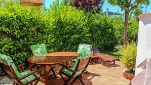 Eine Terrasse mit Holztisch und -stühlen, umgeben von üppigen grünen Büschen und einem kleinen Baum.