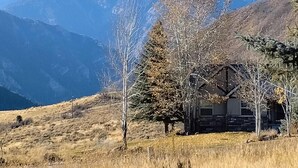 Una baita rustica con facciata in pietra, immersa in un campo erboso e arido con montagne innevate sullo sfondo.