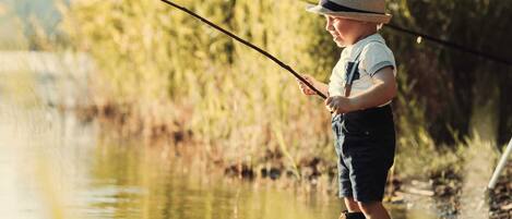 A child fishing by a calm river with greenery in the background.
